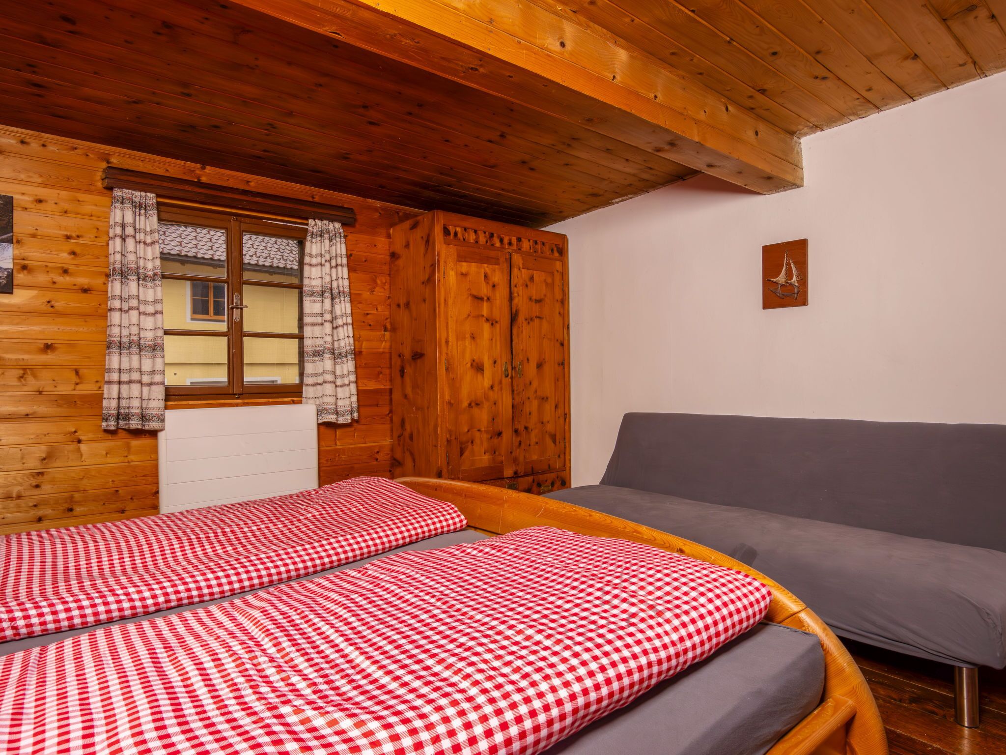 A room with two beds with checkered bedding, a grey sofa, wooden walls, and a window with patterned curtains.