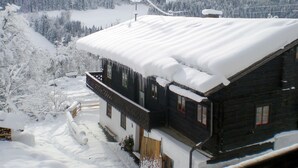 Una cabaña de madera con techo nevado, rodeada de un paisaje invernal y montañas.