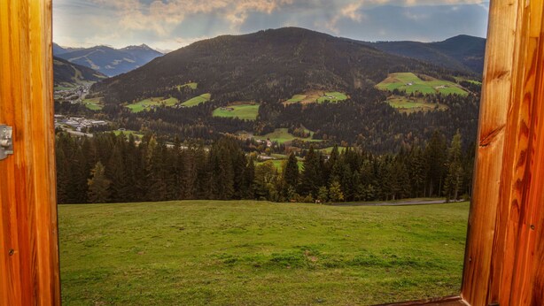 Vue de l’intérieur d’un chalet en bois, donnant sur un paysage montagneux avec des champs verdoyants et une vallée.