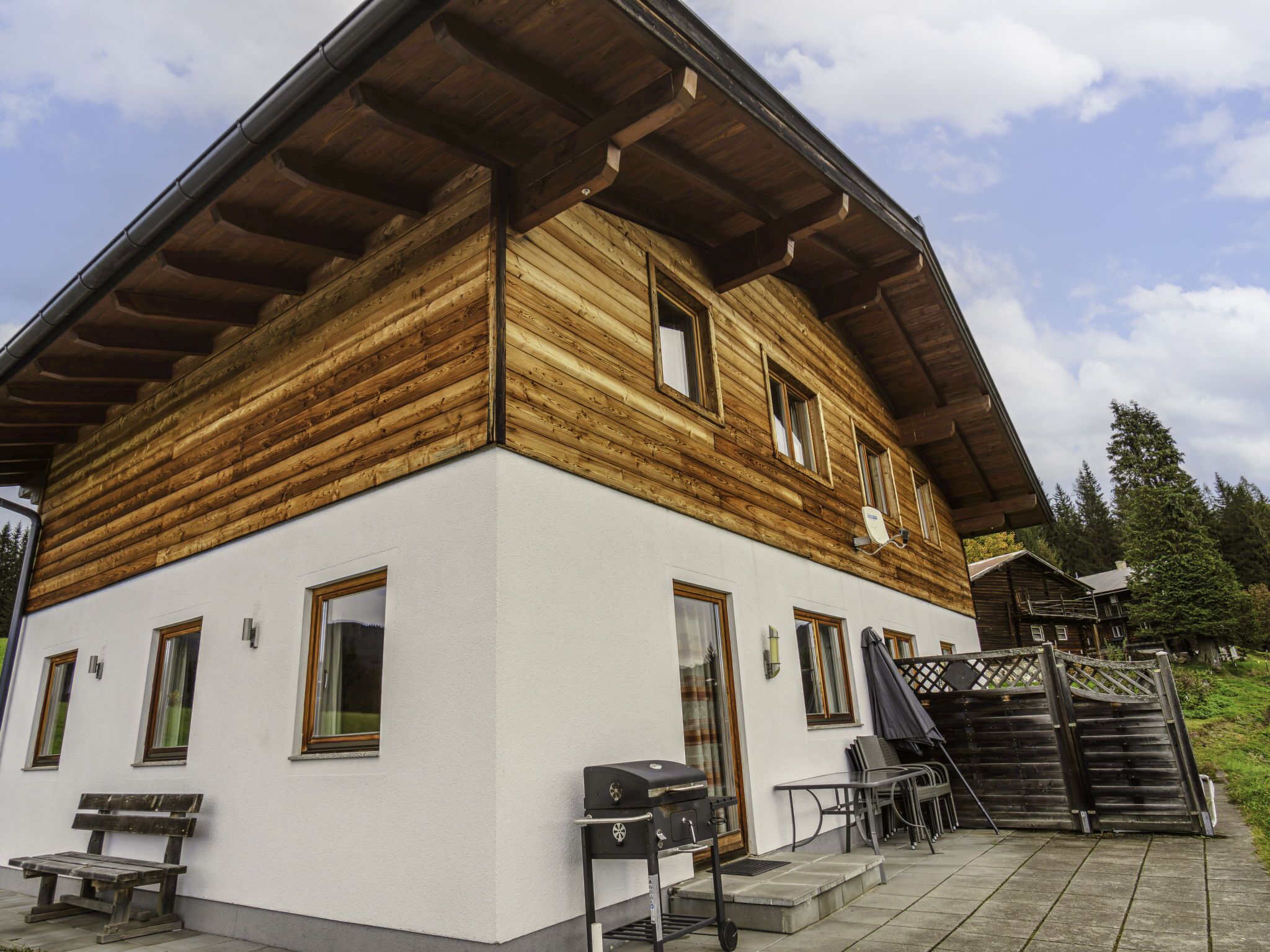 Een houten chalet met een witte gevel, een overdekte veranda en een barbecue buiten.