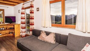 Habitació acollidora amb sostre de fusta, finestra gran amb vista a la muntanya, sofà amb coixins estampats i escriptori de fusta amb televisió.
