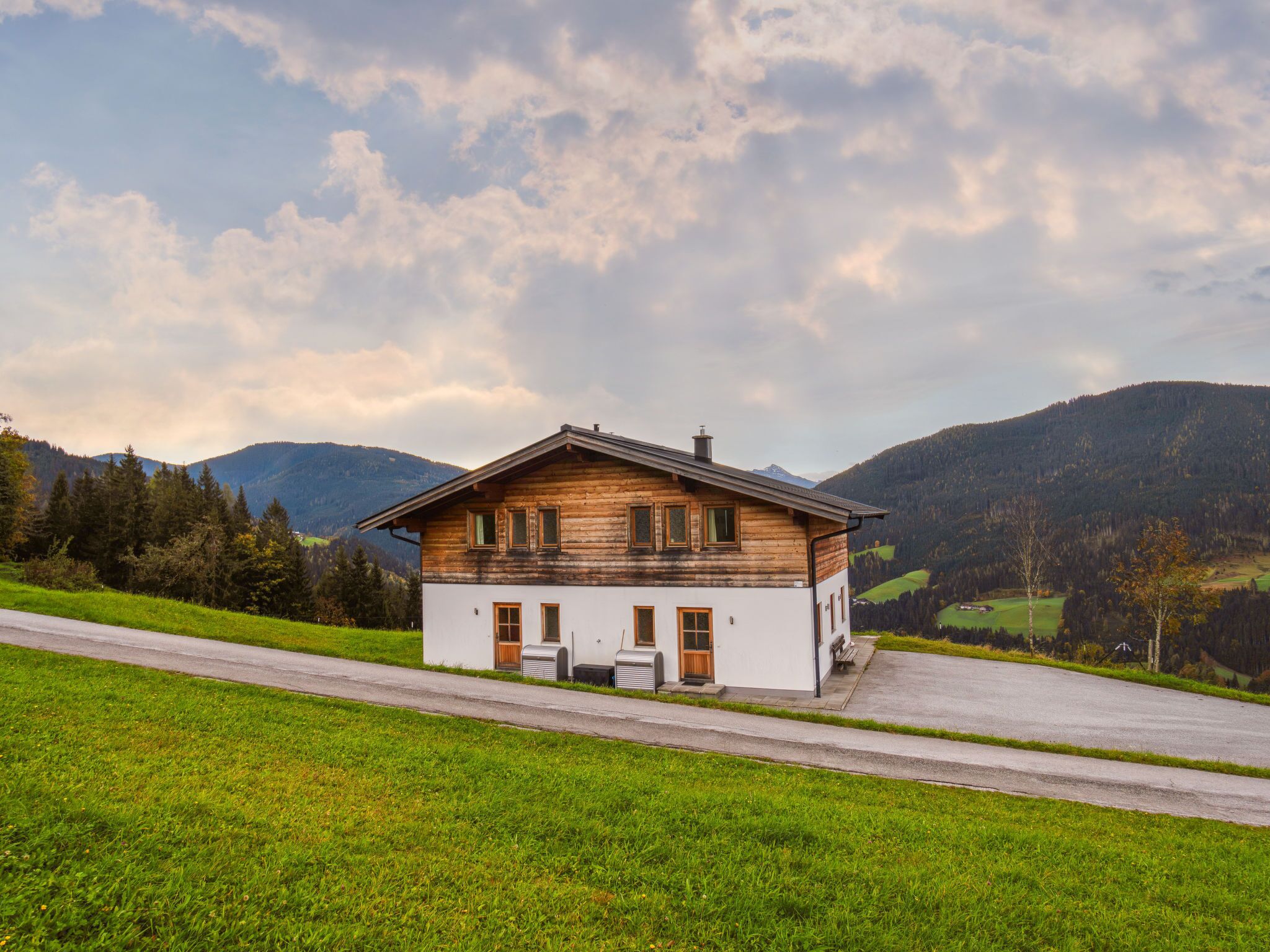A wooden house with a sloped roof, located on a grassy hillside with a road in the foreground and mountains in the background.