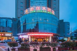 A hotel with a curved facade, illuminated signage, and a paved entrance area with potted plants.