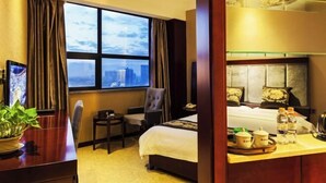 Ein Hotelzimmer mit großem Fenster, einem Bett, einem Schreibtisch mit Pflanze, einem Sessel und einem Essbereich mit Tablett.