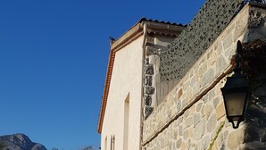 Un bâtiment en pierre avec un toit recouvert de tuiles, une grande plante en pot et un chemin menant à une entrée en arc de cercle.