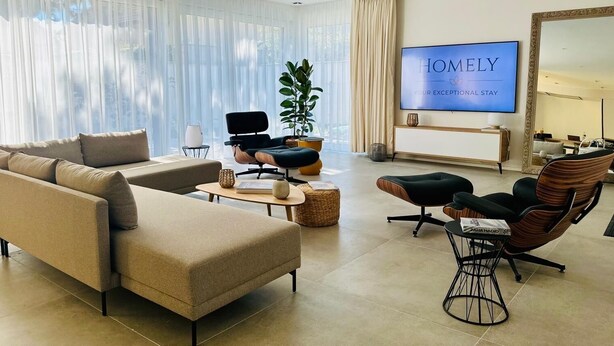 A modern living room with a beige sofa, a black chair, a wooden coffee table, and a TV displaying "HOMELY YOUR EXCEPTIONAL STAY". - Homely Basel Airport (Saint-Louis)