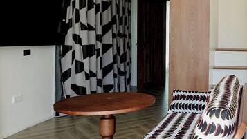 Ein modernes Wohnzimmer mit einem runden Holztisch, einem Korbusofa mit gemusterten Kissen, einem schwarz-weißen geometrischen Vorhang und einer Deckenleuchte.