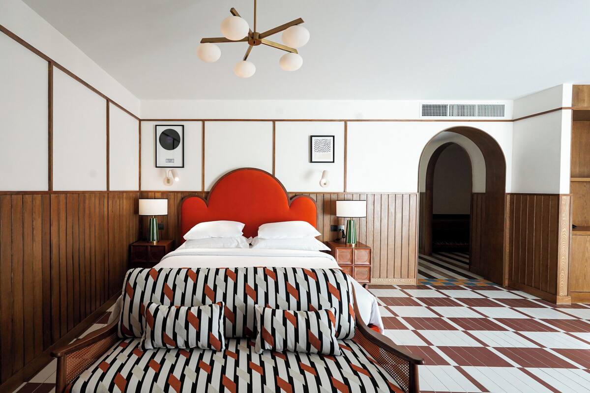 Une chambre d’hôtel avec un grand lit, des boiseries, une tête de lit rembourrée en tissu rouge, des coussins à motifs, un ventilateur de plafond et un sol à carreaux.