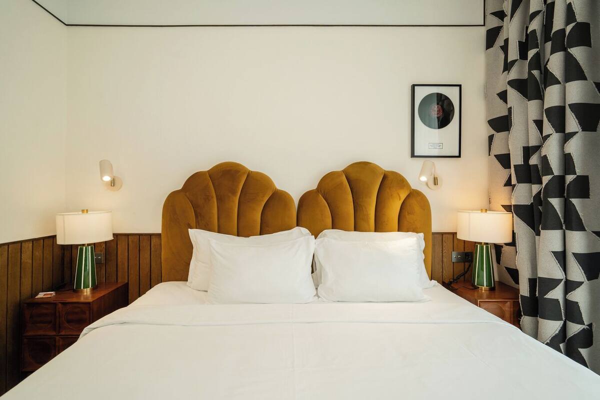 Un lit doté d’une tête de lit originale, deux tables de chevet avec des lampes, un tableau encadré au mur et un rideau à motifs.
