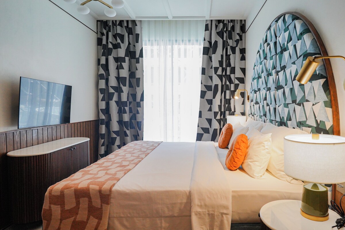 Une chambre d’hôtel avec un lit, une table de chevet, une lampe, une télévision et des rideaux à motifs.