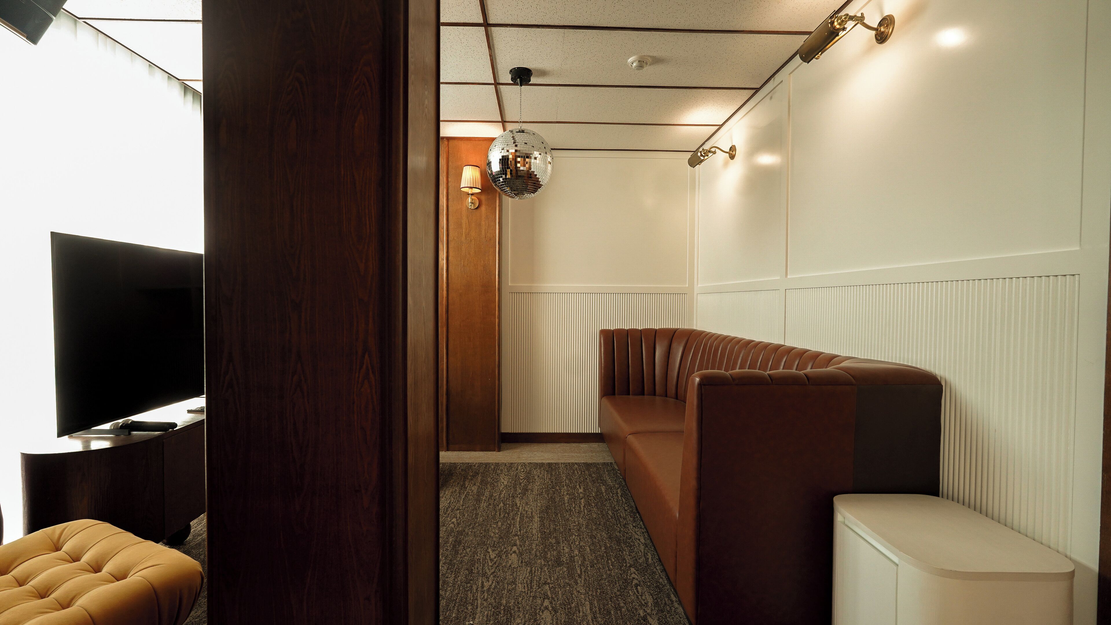 Een moderne hotelkamer met een groot flatscreen televisietoestel, een bruine leren bank, een klein rond tafeltje en een houten deur.