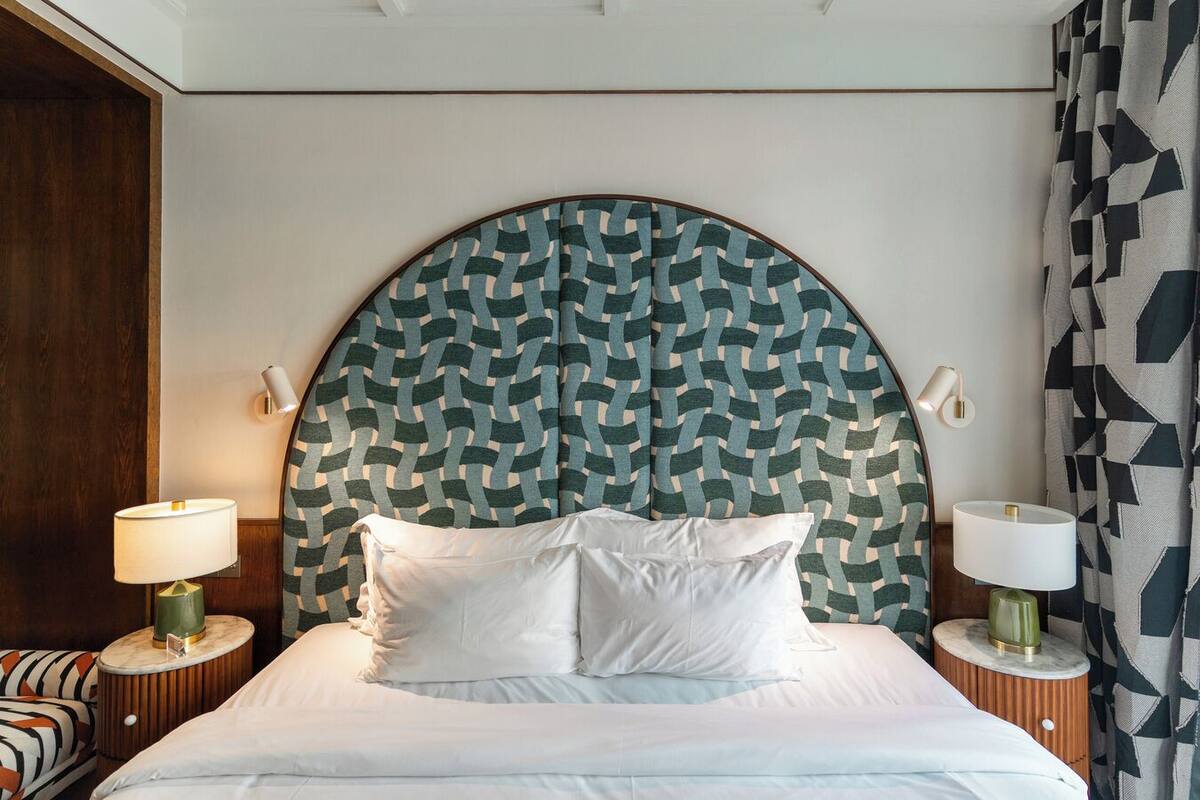 Une chambre d’hôtel avec un grand lit, deux tables de chevet avec des lampes, une tête de lit à motifs géométriques et un rideau à motifs.