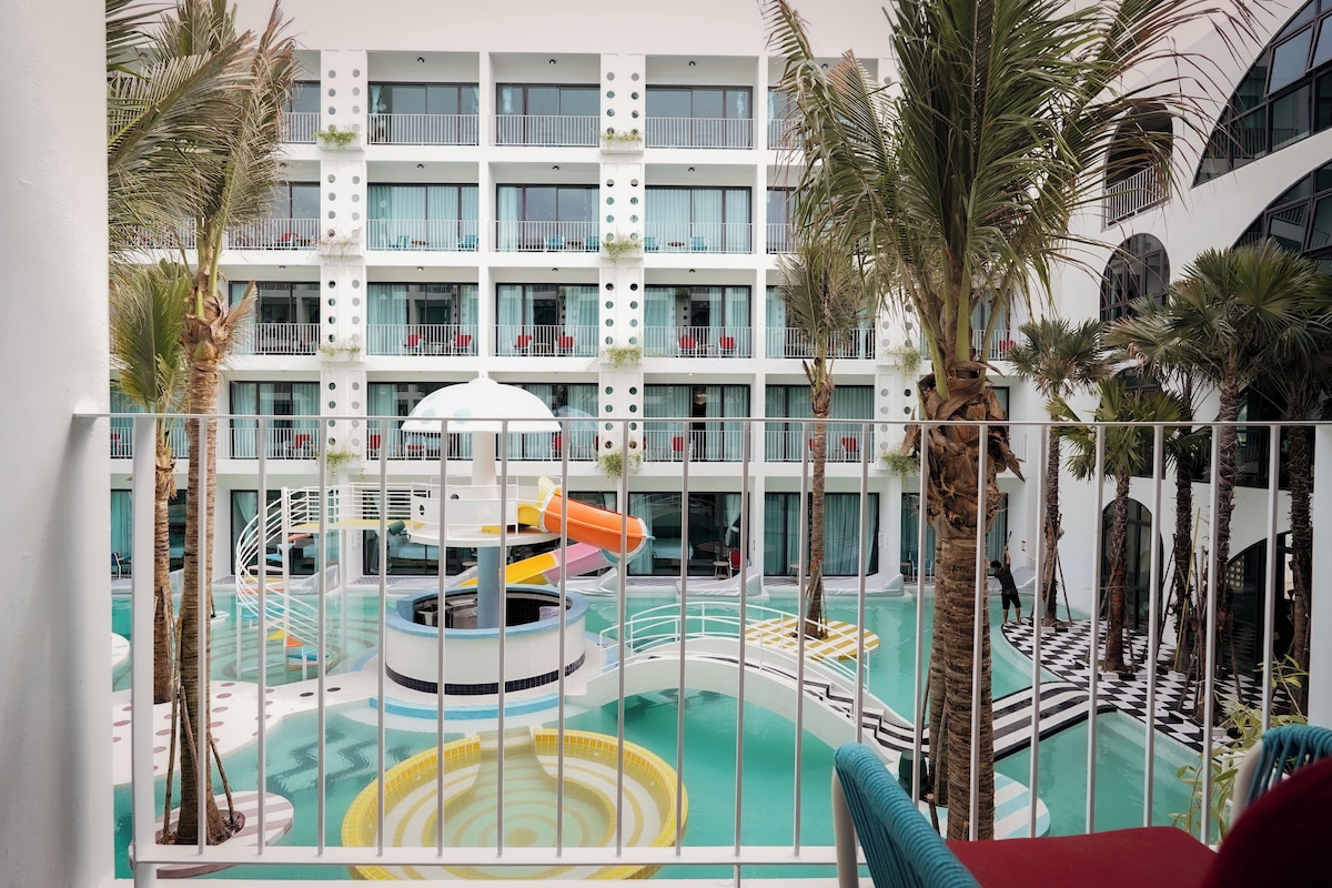 Uma área de piscina em vários níveis, com um toboágua, rodeada por palmeiras e um edifício branco com varandas.