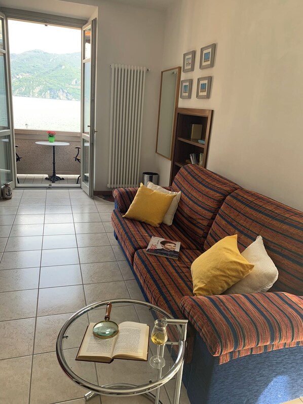 Un salon avec un canapé, une petite table sur laquelle se trouve un livre et un verre de vin, un radiateur et une vue sur l’extérieur.