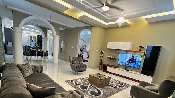 A spacious living room with a large sectional sofa, a smaller armchair, and a coffee table. There is a flat-screen TV mounted on the wall, a chandelier, and a decorative rug. - Nanani Luxury Villa - Kumasi (Kumasi)