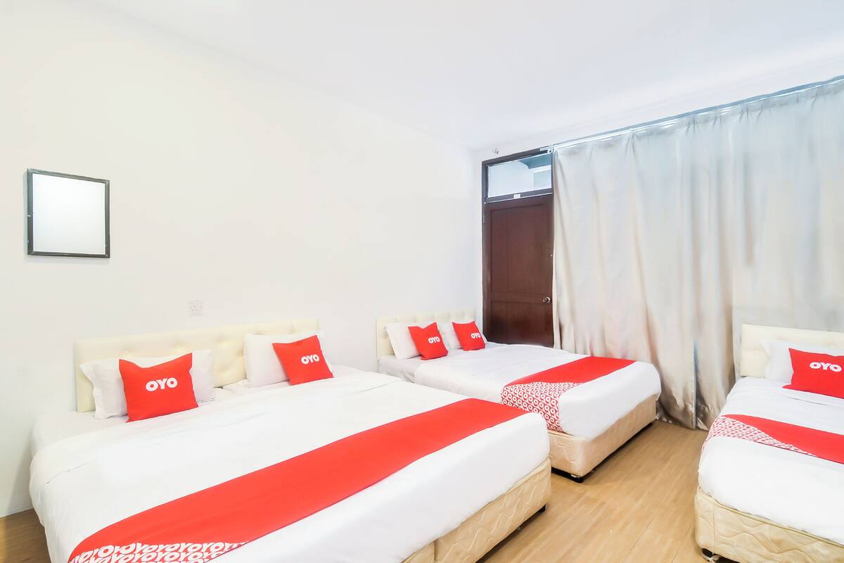 Une chambre d’hôtel avec trois lits, chacun agrémenté de draps blancs et de touches de couleur rouge. On aperçoit une fenêtre avec des rideaux blancs et une porte.