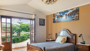 Un dormitorio con una cama grande, una mesita de noche, una lámpara, un cuadro y vistas al verdor a través de un ventanal abierto.