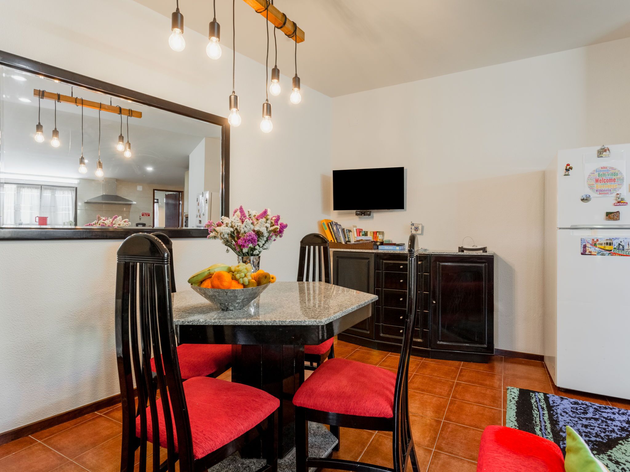 A modern kitchen with a dining area, featuring a granite countertop, red upholstered chairs, a bowl of fruit, and a flat-screen TV mounted on the wall.