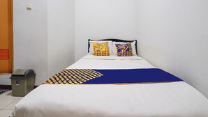 Un lit bien fait, recouvert d’une couvre-lit aux motifs bleus et jaunes, avec deux oreillers aux motifs géométriques, et une petite poubelle placée à gauche.