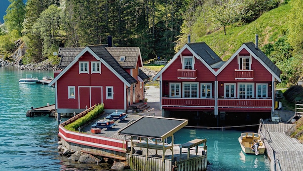 Ein rotes Holzhaus mit einem Steg, einem Boot und einem bewaldeten Hügel.