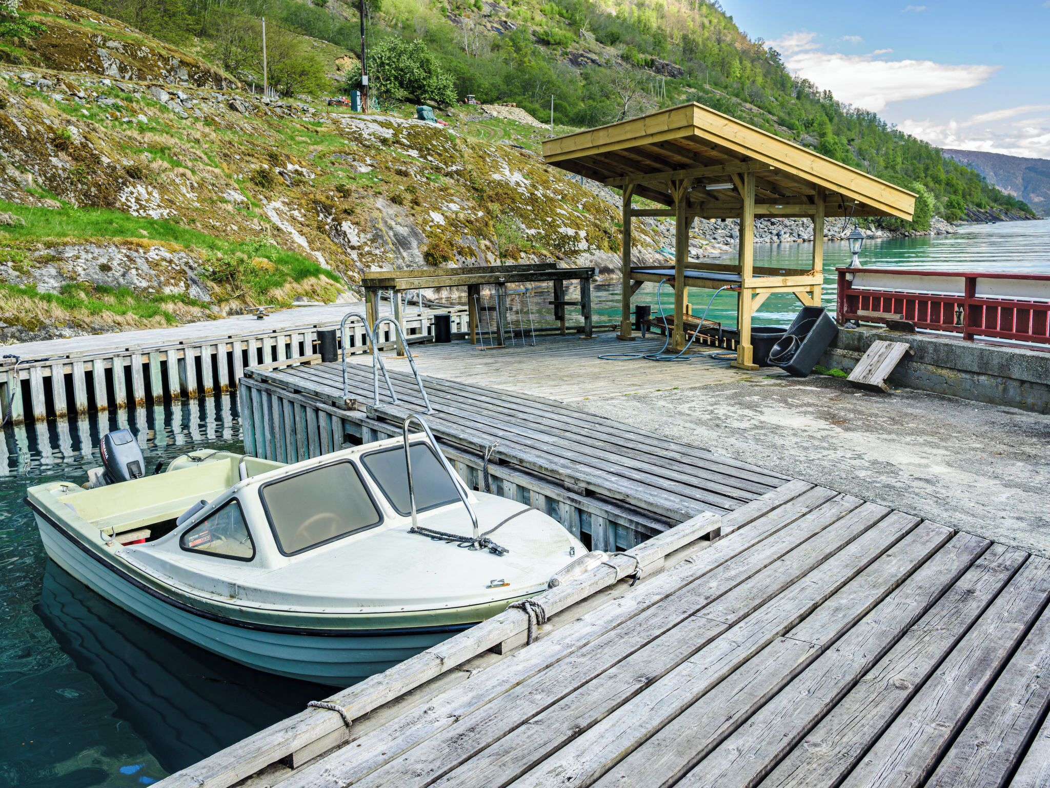 Une jetée en bois avec un bateau amarré au quai, un espace de repos abrité et, en arrière-plan, une colline rocheuse.