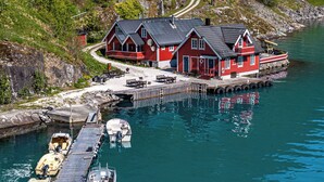 Ein rotes Haus mit einem Steg, der sich in ruhiges Wasser erstreckt, Boote liegen am Steg festgemacht, und ein Weg führt zum Haus.