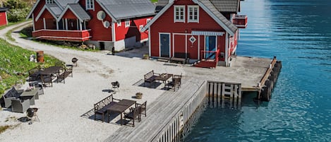 A wooden pier leading to a red building with a dock, outdoor seating, and a view of a lake.