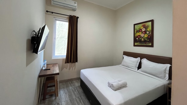 A hotel room with a bed, a desk, a window with curtains, an air conditioner, and a painting on the wall.