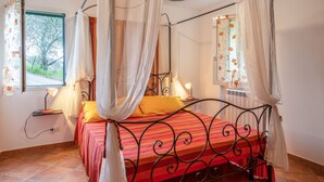 Un lit à baldaquin avec une couvre-lit à rayures rouges et oranges, des rideaux blancs et une vue sur un arbre par la fenêtre.