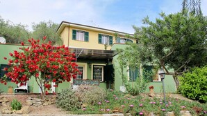 Une maison à deux étages, avec un mur de pierre à l’avant, un arbre à fleurs rouges et un jardin agrémenté de fleurs roses.
