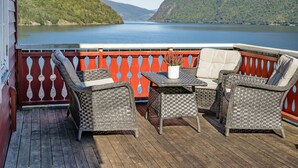 Um terraço com móveis de vime e um vaso de planta, com vista para um lago e montanhas ao fundo.