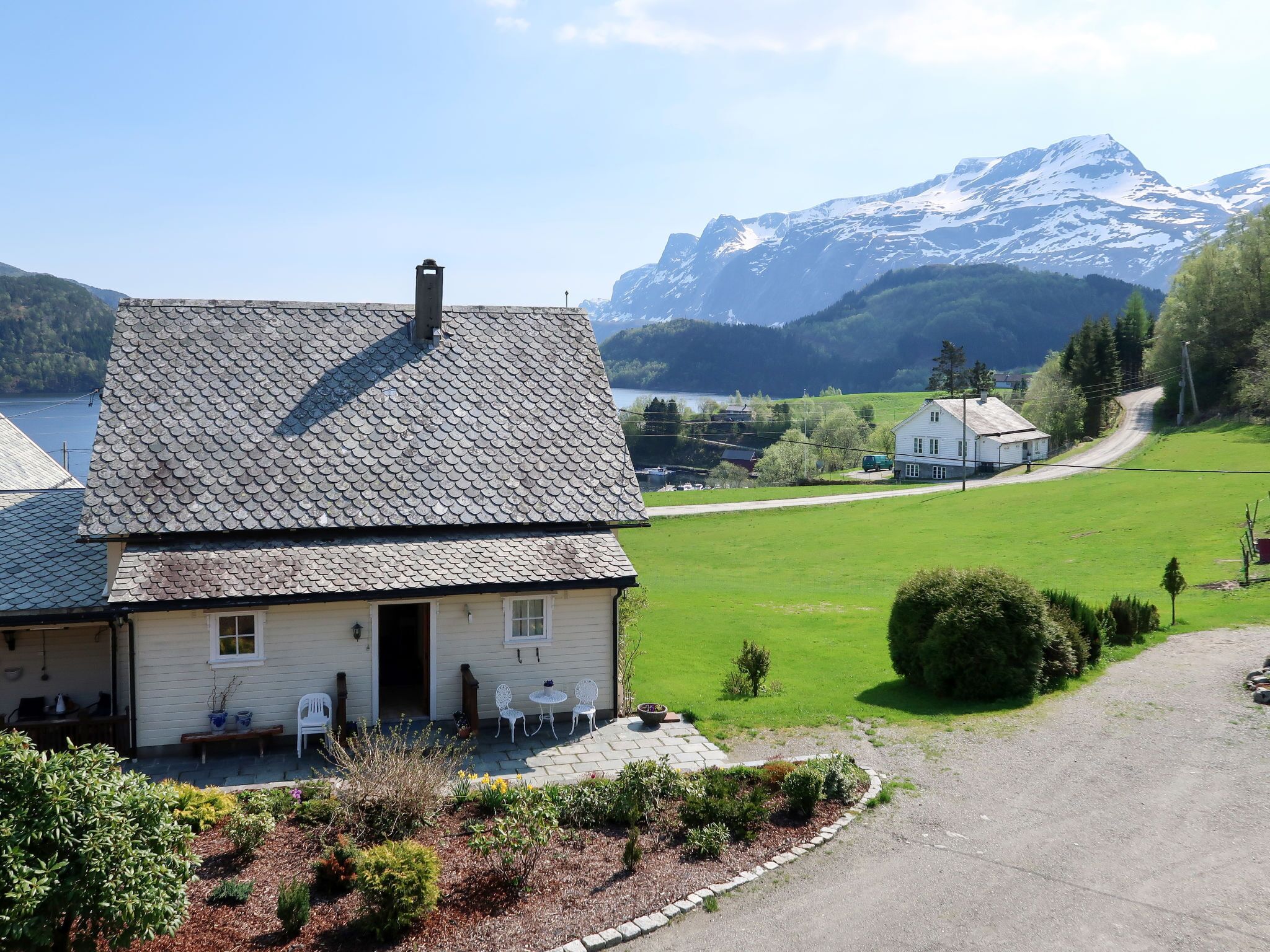 Ein Haus mit schrägem Dach und Schornstein, einer gepflasterten Auffahrt und einem gepflegten Garten mit Bank und Stühlen.