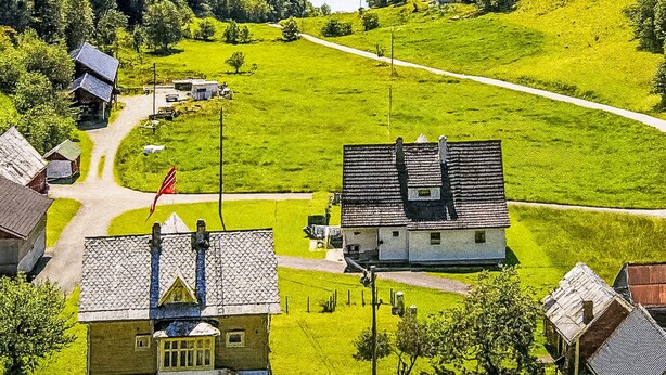 Un paysage rural avec un vaste champ ouvert, quelques bâtiments et une étendue d’eau en arrière-plan.