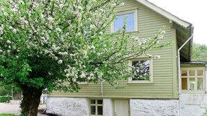 Une maison à l’extérieur vert et aux bordures blanches, un grand arbre aux fleurs blanches et une petite terrasse avec des chaises en osier.