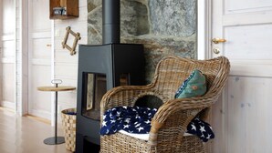 A cozy room with a stone wall, a black wood-burning stove, a wicker chair with a blanket, and a small wooden shelf with a clock. - Elistova by Interhome (Onarheim)