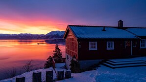 A cabin with a snow-covered roof, a wooden deck, and a view of a calm lake at sunset. - Elistova by Interhome (Onarheim)