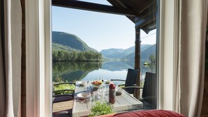 A lakeside view from a room with a table set for two, featuring a glass of water, a bowl of fruit, and a bottle of sauce.