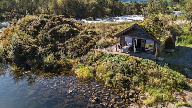 A rustic cabin by a river with a clear view of mountains in the background.