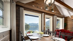 A dining area with a wooden table set for four, large windows offering a view of mountains and a lake, and a chandelier above.