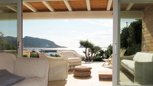 Un salon moderne avec un canapé blanc, des fauteuils en osier et une porte vitrée donnant sur un balcon d’où l’on peut admirer la mer et les montagnes.