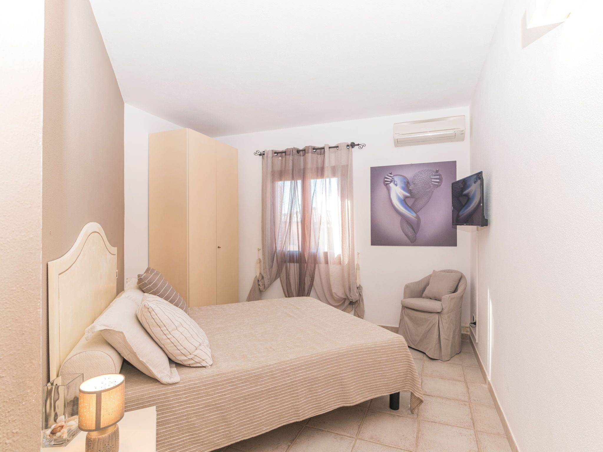 A bedroom with a bed, a bedside lamp, a TV mounted on the wall, and a window with curtains.