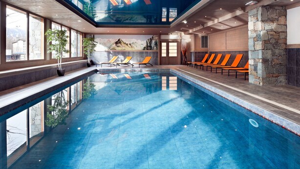 Une piscine intérieure à l’eau d’un bleu cristallin, entourée de chaises longues et d’un mur en pierre.