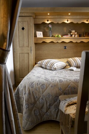 Un dormitorio acogedor con cabecera de madera, cama con ropa de cama floral, mesita de noche con lámpara y estante con objetos decorativos.