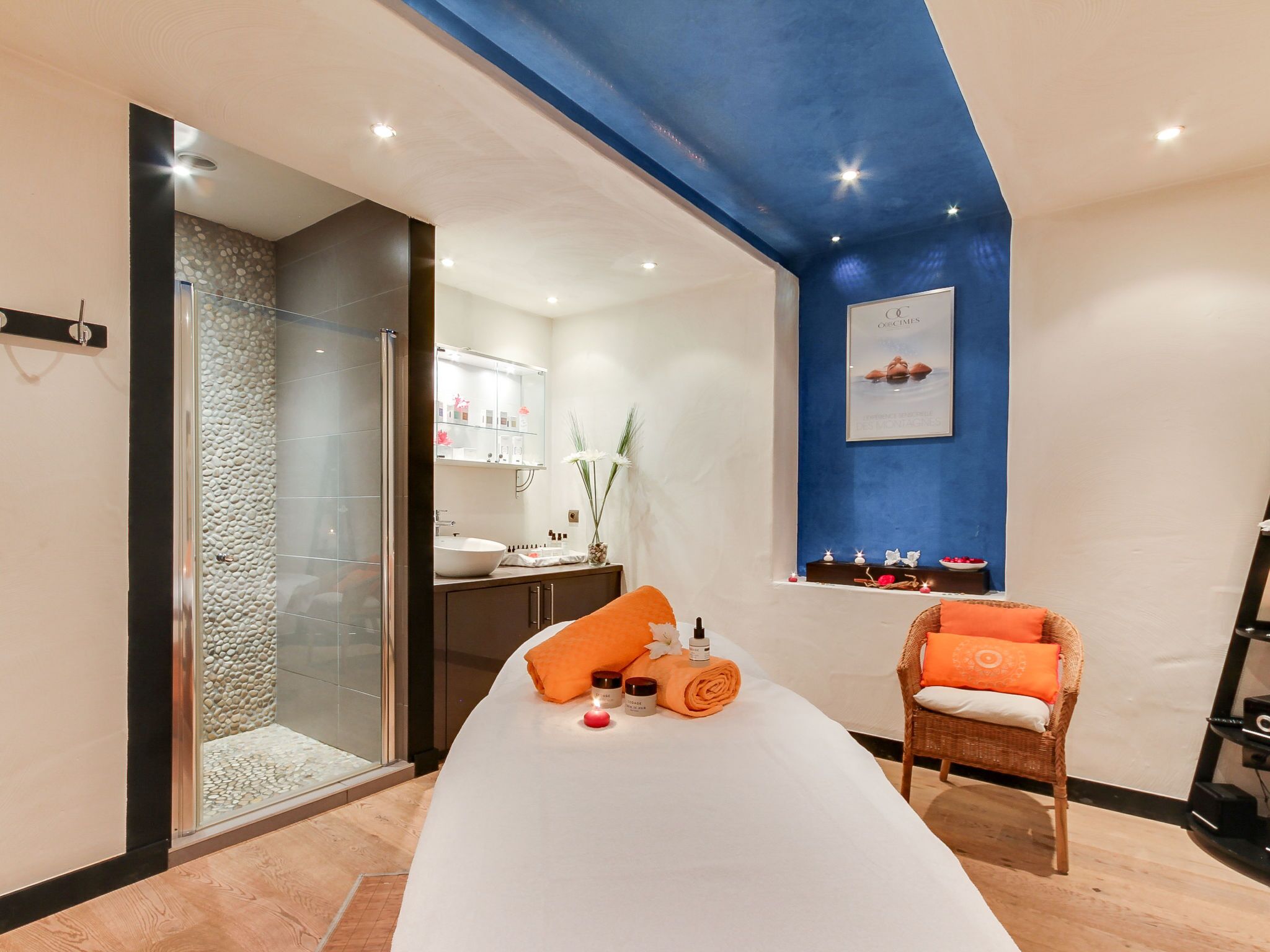 Ein moderner Wellnessraum mit einer Duschkabine aus Glas, einem weißen Bett mit orangefarbenen Handtüchern, einem Korbstuhl und einer blauen Akzentwand.
