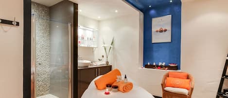 Ein moderner Wellnessraum mit einer Duschkabine aus Glas, einem weißen Bett mit orangefarbenen Handtüchern, einem Korbstuhl und einer blauen Akzentwand.