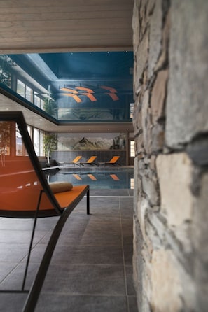 Un área de piscina interior moderna con pared de piedra, sillones de descanso y vistas a un paisaje montañoso a través de un techo de cristal.