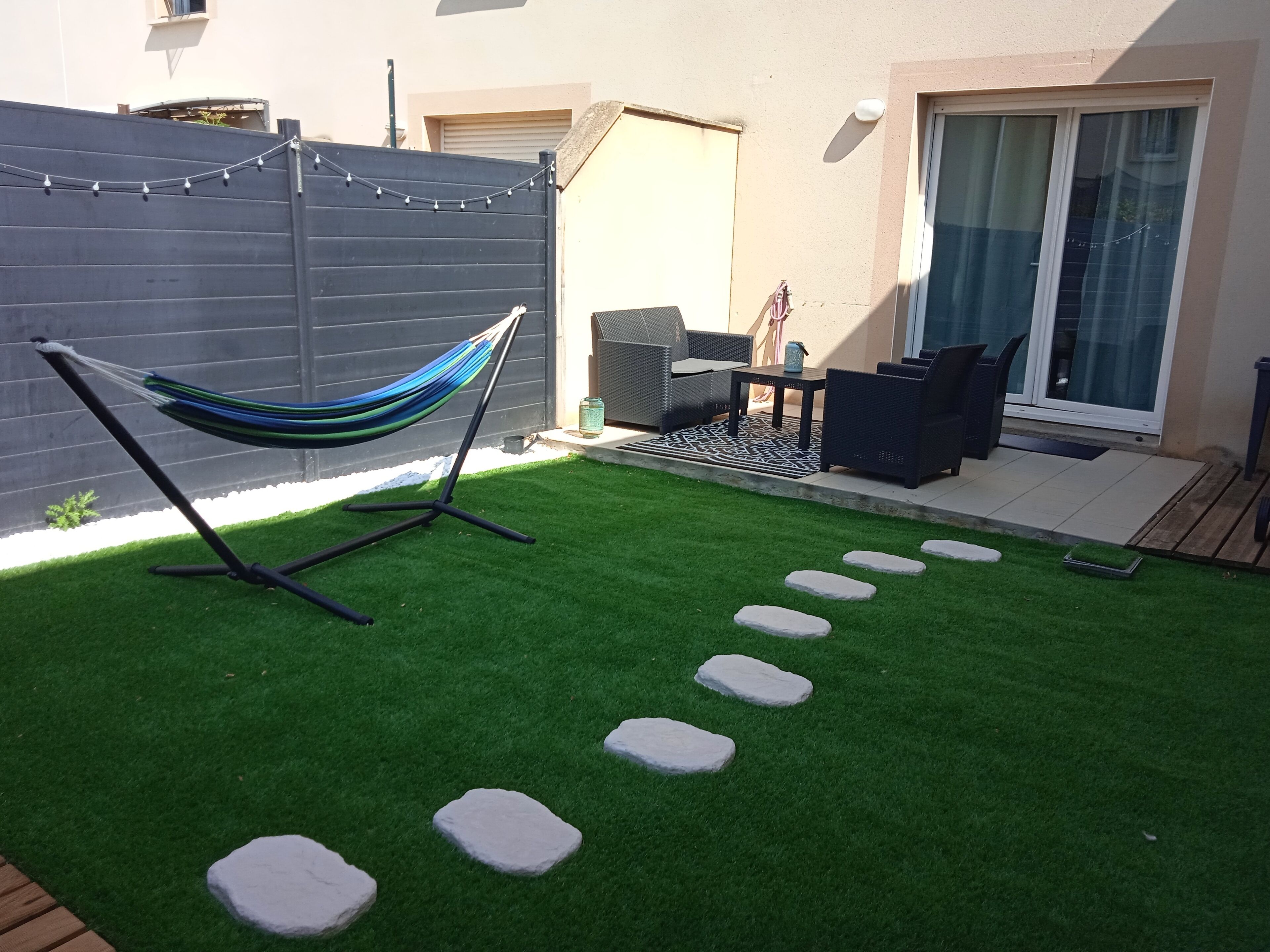Un patio trasero con césped artificial verde, una hamaca, mobiliario exterior y una zona de patio con puerta corrediza de vidrio.