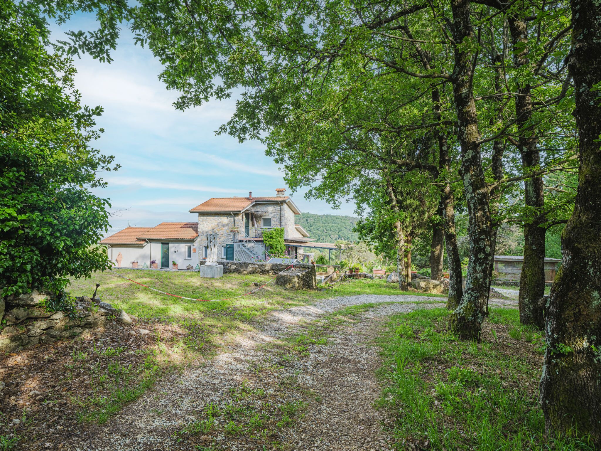 Une maison en pierre avec un toit recouvert de tuiles rouges, entourée d’arbres et bordée d’un chemin de gravier.