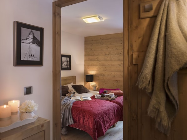 A cozy bedroom with a wooden headboard, a bed with a red quilt, a bedside table with candles, and a framed picture on the wall.