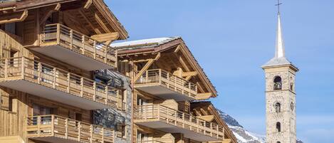 A wooden chalet with balconies, a stone bell tower, and a snowy mountain backdrop.